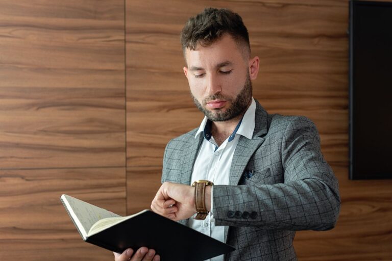 Young businessman in gray suit checking wristwatch while holding a notebook indoors.